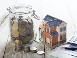 Une petite maquette de maison posée sur une table entre une calculette et un pot en verre rempli de pièce de monnaies.