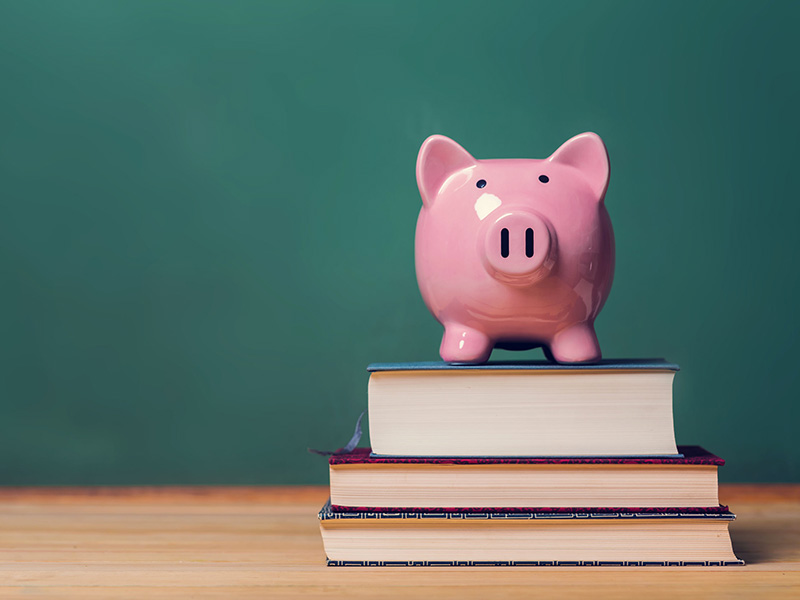 Une tirelire et des livres placés sur une table, devant un tableau vert d'école.