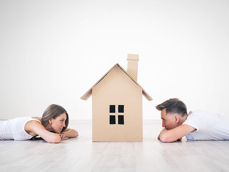 Jeune couple couché sur le ventre et se faisant face, avec, au centre, une maison de carton.