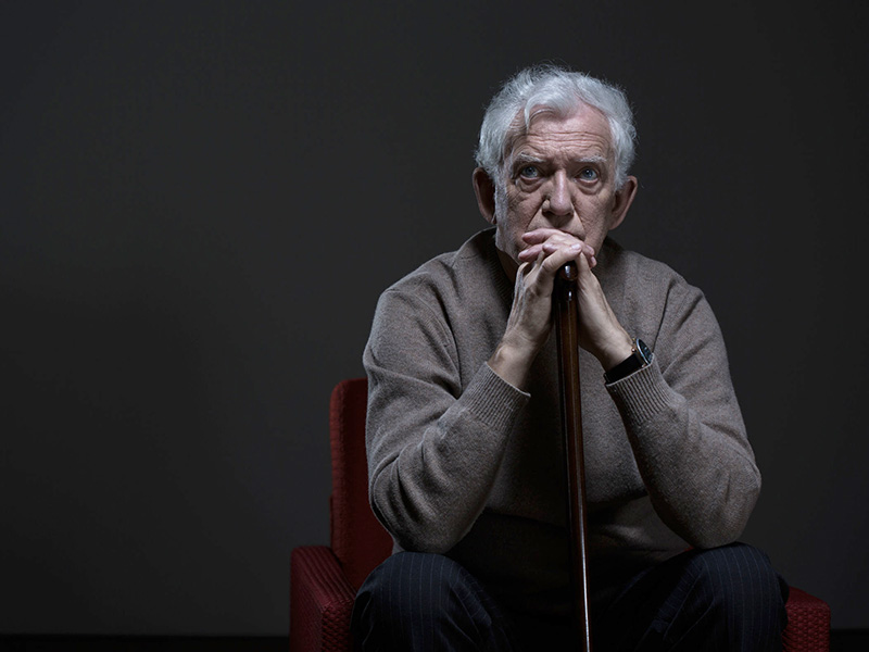 Homme âgé, pensif, assis sur une chaise dans la pénombre.