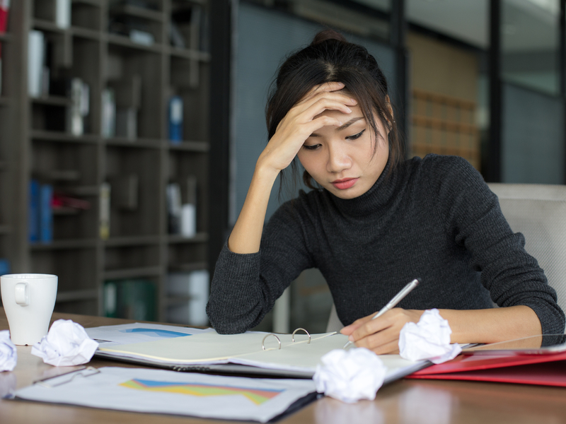 Une femme devant un bureau avec un stylo. Des papiers sont devant elles, certains sont froissés. Elle semble soucieuse.