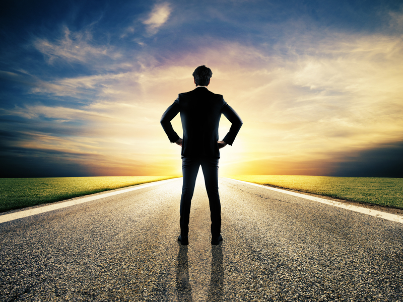 Un homme d'affaire debout sur une route, les mains sur les hanches face à un lever de soleil.