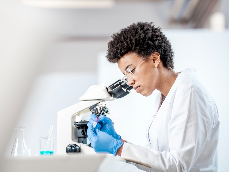 Une femme habillée en médecin étiquetant un tube à essai. Sur la table à côté d'elle on voit un microscope et des fioles.