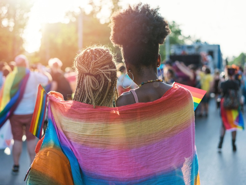 Deux jeunes femmes qui marchent en se tenant par la taille. Derrière elle, un drapeau de la Fierté.