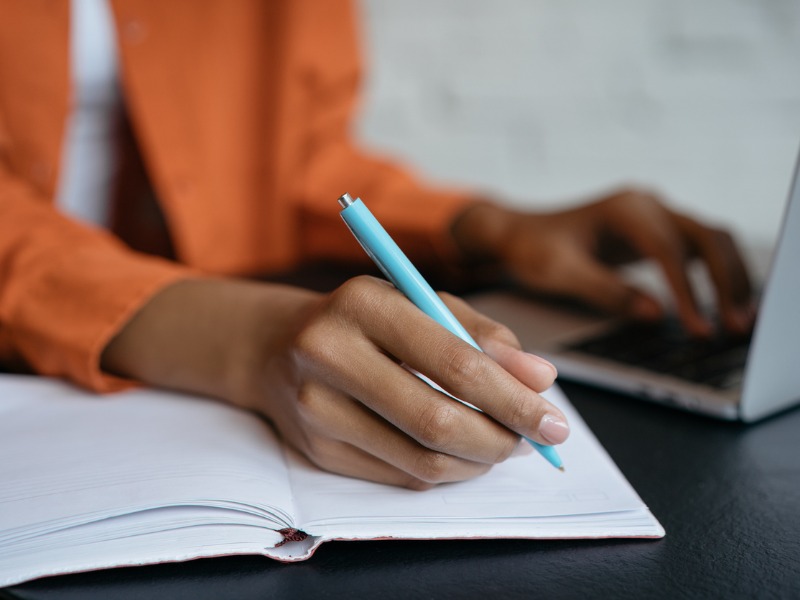 Une femme assise à une table devant son ordinateur. Elle écrit dans un cahier en regardant son écran.